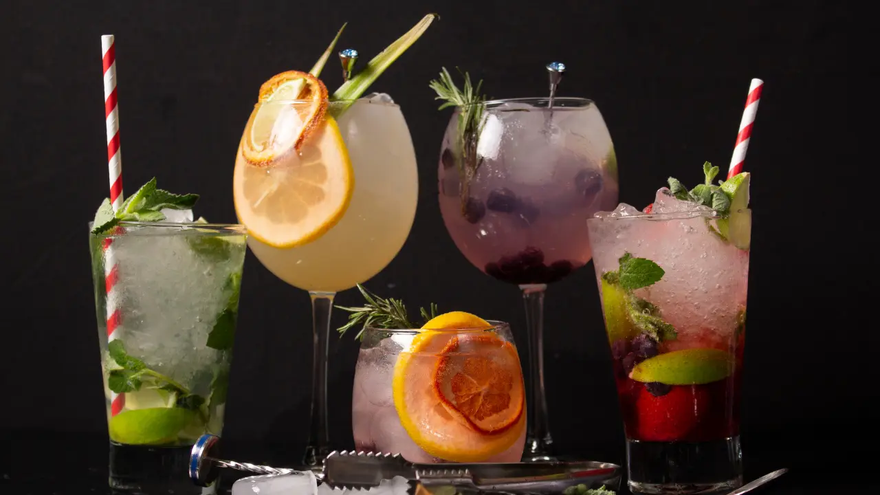 A collection of five colorful cocktails in various glasses. Each drink is garnished with fruit, herbs, and ice.