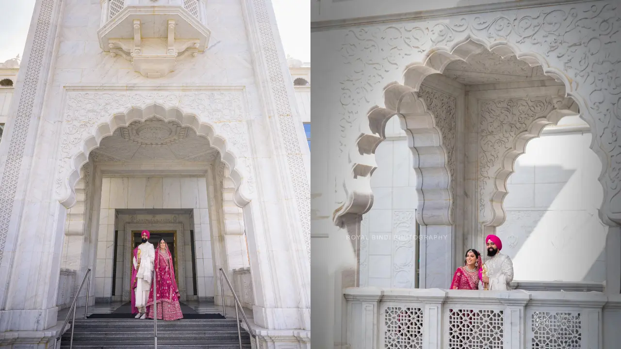 A couple stands under a stunning white archway with intricate carvings. The bride is wearing a pink and gold dress, and the groom is wearing a white turban and white clothes. The couple is smiling and looking at each other. The background is a white marble wall with a beautiful archway and a balcony. The image is taken from a low angle, giving the viewer a sense of grandeur.