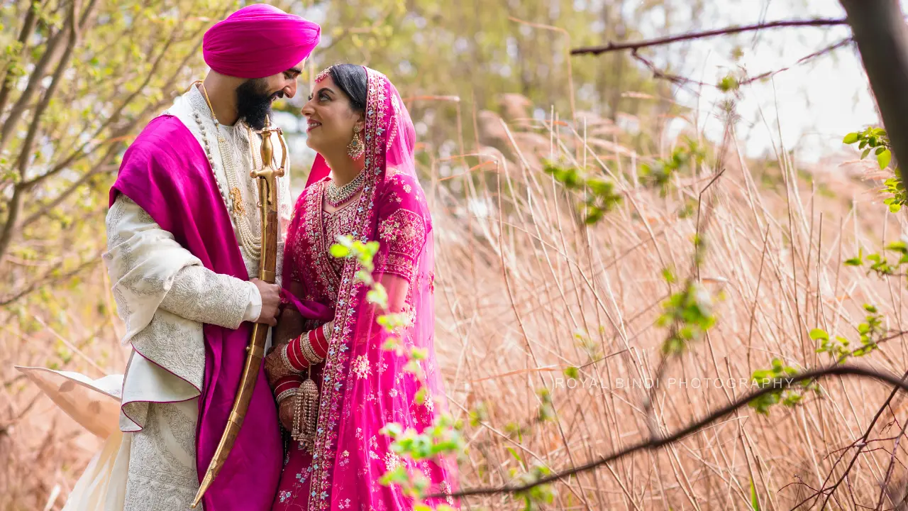 A Sikh couple in traditional clothing standing in a field of tall grass, looking at each other with love.