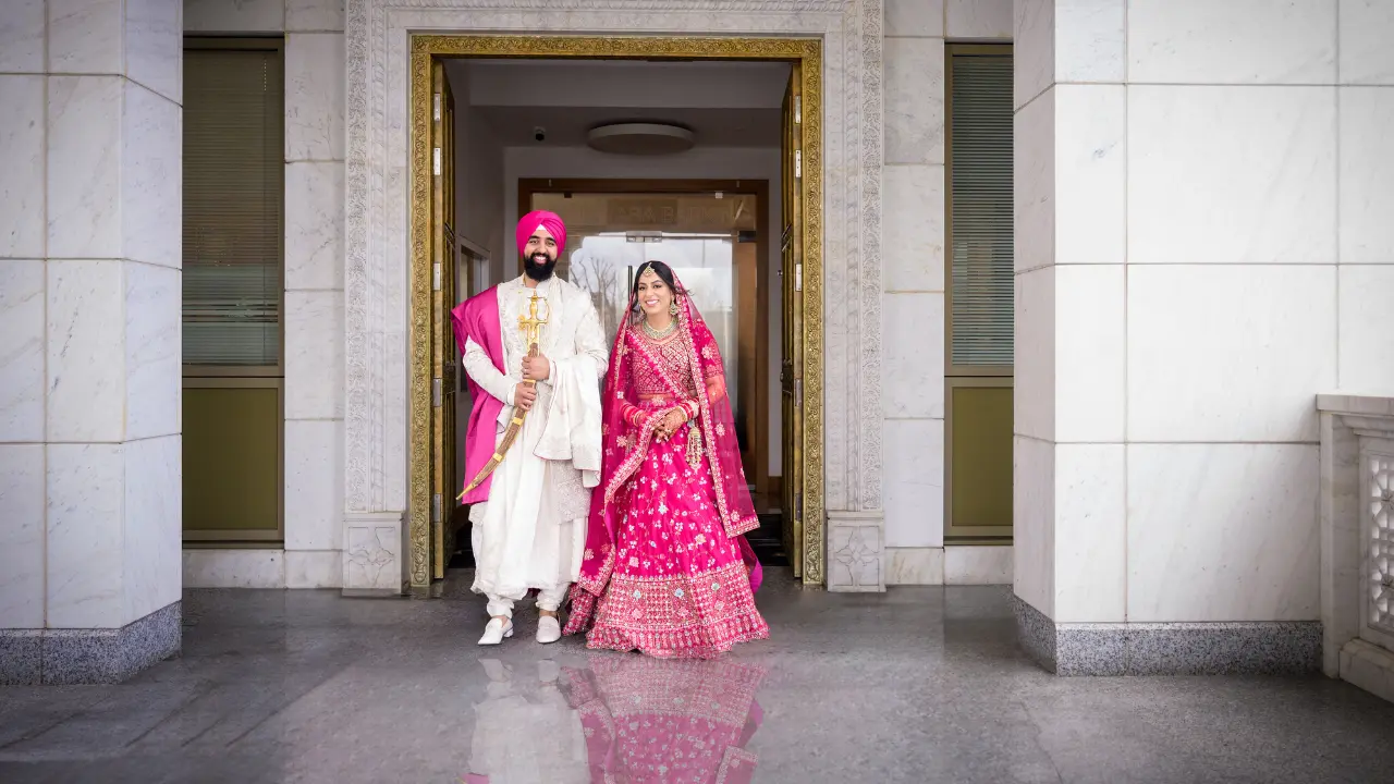 A newly married Sikh couple poses for a photo outside a Barking Gurdwara's building. The bride is wearing a bright pink lengha, and the groom is wearing a white turban and a long white robe. They are both smiling and looking at the camera.