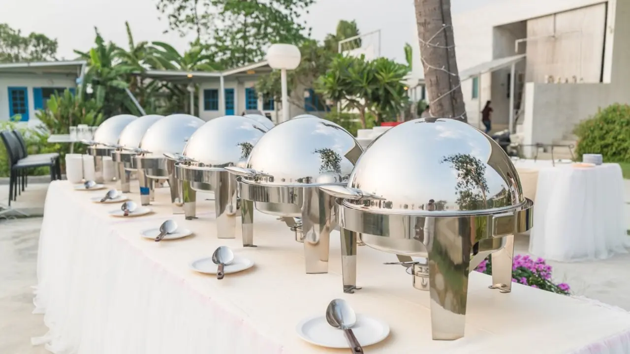 A row of silver chafing dishes set up on a white tablecloth outdoors, ready for a catered event.