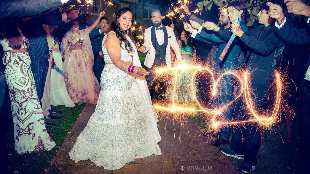 A newly married couple stands in front of their wedding guests who are holding sparklers. The sparklers are creating the words "I love you" in the air. The bride is wearing a white wedding dress and the groom is wearing a black tuxedo. The couple is smiling and looking at each other. The wedding party is celebrating the new couple.