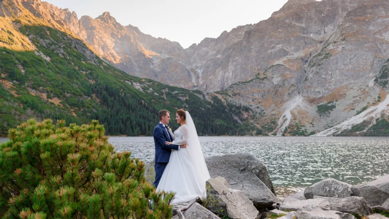 A newly married couple stands by a mountain lake, surrounded by rocks and lush green trees. The bride is wearing a white wedding dress and the groom is wearing a blue suit. They are looking at each other with love and affection. The mountains are in the background, with a clear blue sky above. This is a beautiful and romantic scene, capturing the love and happiness of this special day.