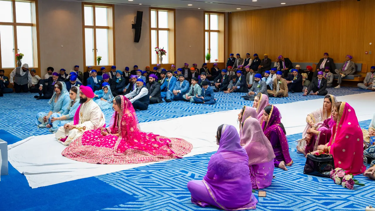 A Sikh wedding ceremony is taking place inside a large Gurdwara. The bride and groom are sitting in the center of the room surrounded by their guests. The bride is wearing a bright pink and gold dress and the groom is wearing a white and gold turban and outfit. The guests are all dressed in their finest clothes and they are smiling and watching the ceremony.