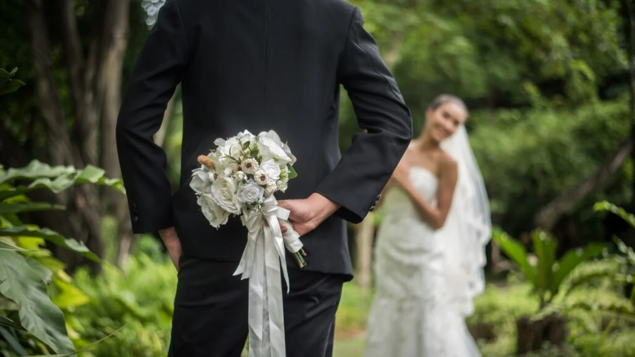 A groom hides a bouquet of white flowers behind his back, smiling at the bride who is walking towards him.