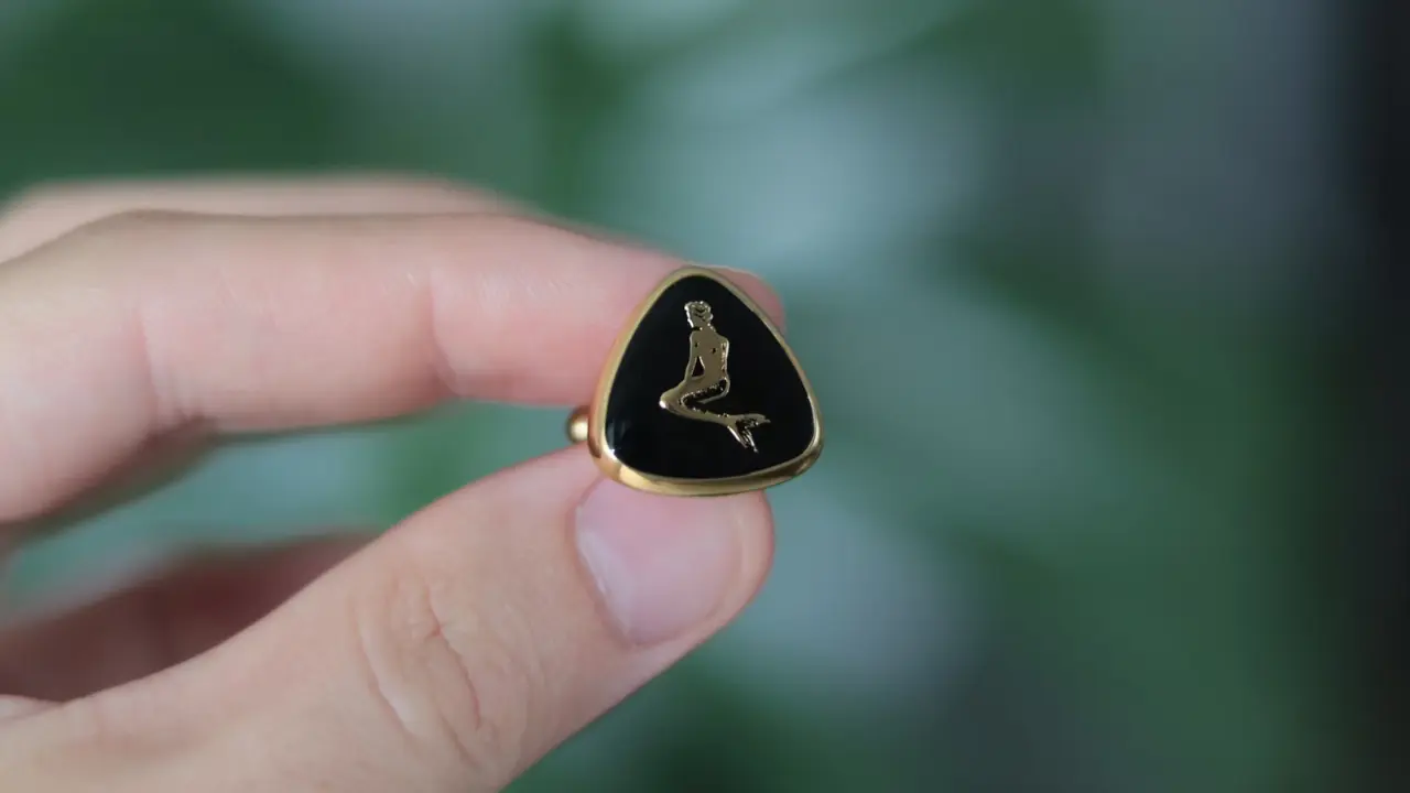 A hand holding a vintage gold ring with a black cufflink featuring a mermaid design.