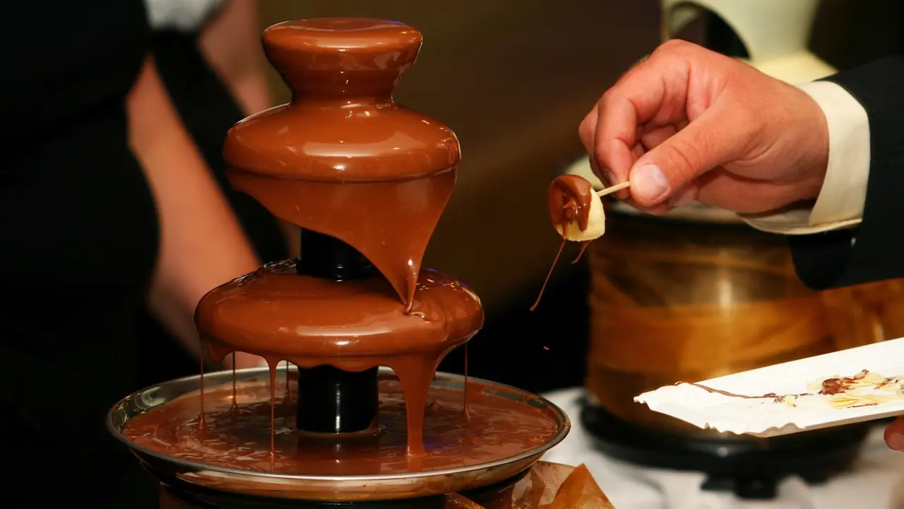 A chocolate fountain with melted chocolate cascading down its tiers. A hand is dipping a banana slice into the chocolate.