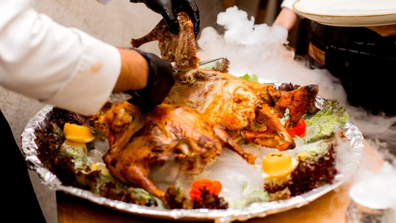 A chef in black gloves carving a roasted bird on a platter surrounded by dry ice smoke.