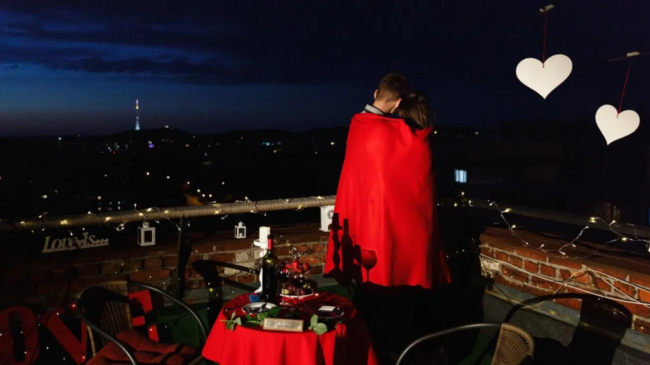 A couple stands on a rooftop with a city skyline in the background. They are wrapped in a red blanket and holding each other close. The table is set with a red tablecloth, a bottle of wine, glasses and two chairs. The setting is romantic and cozy. There are white paper hearts hanging in the air. There is a string of white Christmas lights running along the edge of the building, adding a touch of magic to the scene.