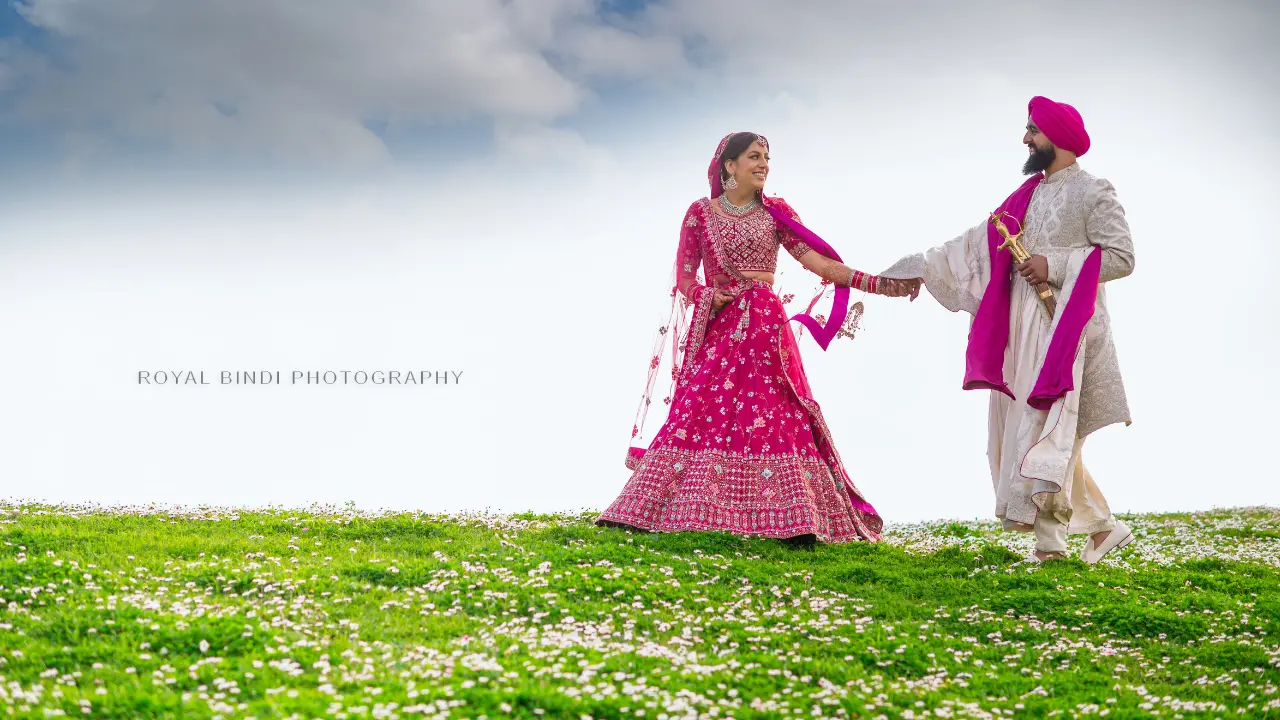 A couple in traditional Indian wedding attire walks hand in hand through a field of flowers. The bride is wearing a pink lehenga and the groom is wearing a white sherwani. They are looking at each other with smiles on their faces. The image is captured by Royal Bindi Photography.