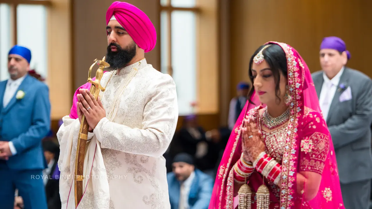 A Sikh groom stands holding a ceremonial sword while the bride stands next to him praying with their hands together. They are both wearing traditional clothing. The ceremony is taking place in a large room.