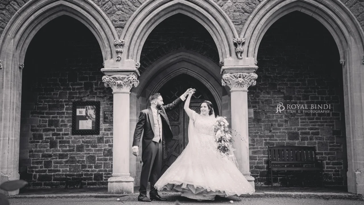 A black and white photo of a wedding couple dancing under arches. The groom is wearing a suit and the bride is wearing a wedding dress. The photographer's logo is in the bottom right corner.