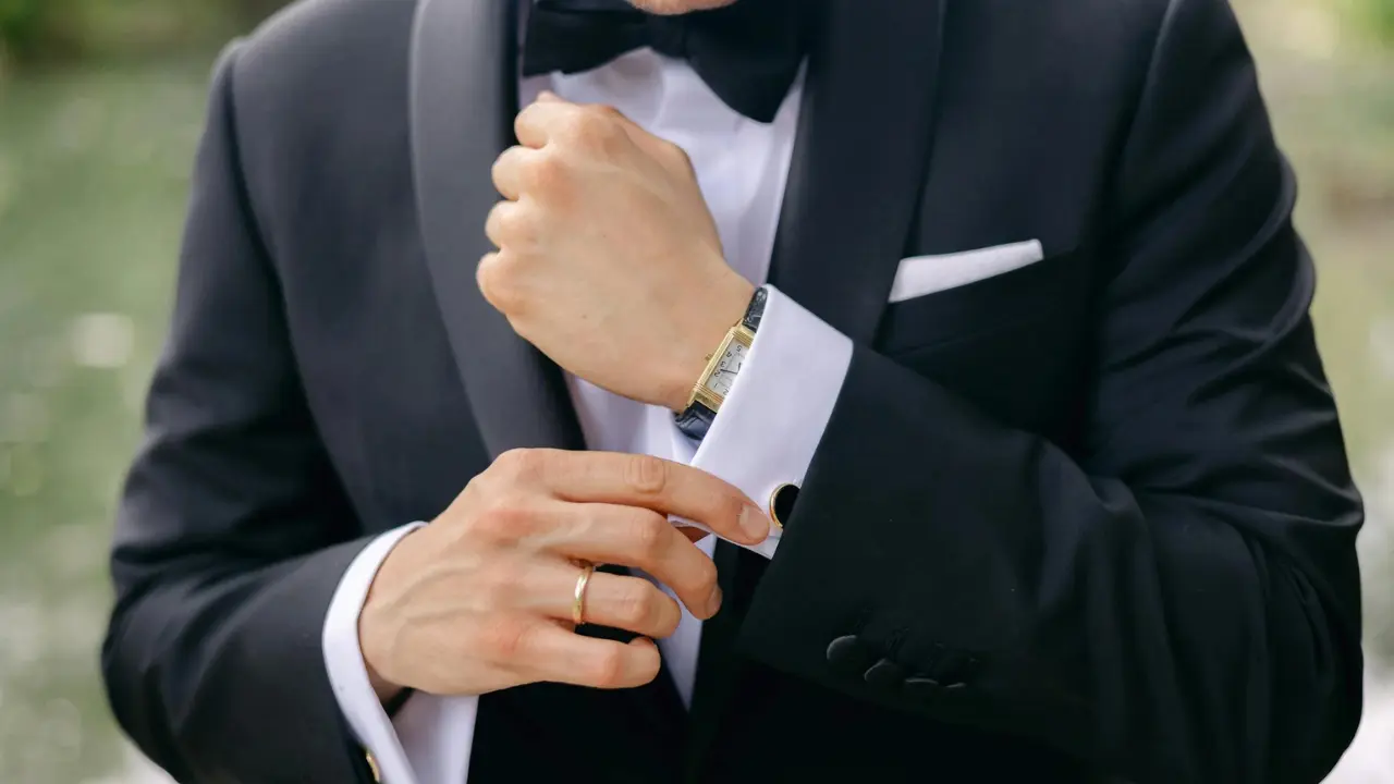 A man is adjusting his cufflinks on his tuxedo suit. He has a classic black bowtie and is wearing a gold watch. He is wearing his engagement ring.