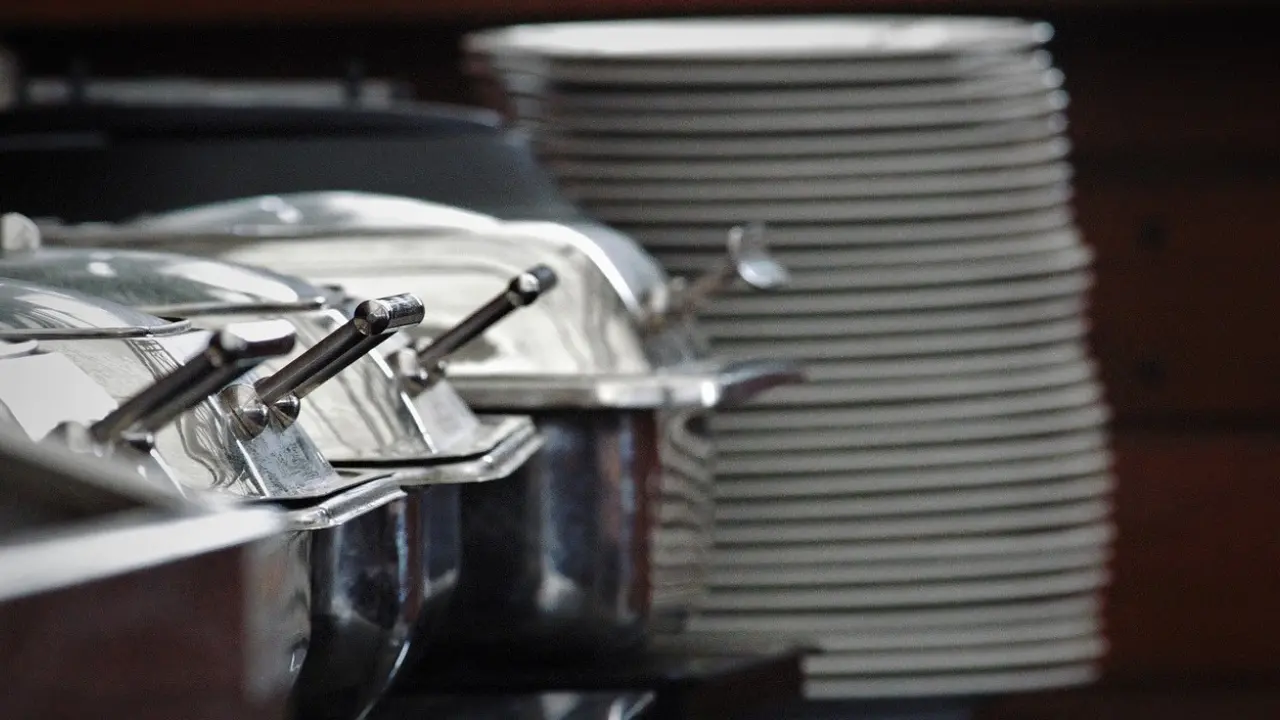 A row of silver chafing dishes with lids and handles, ready to serve food. A stack of white plates is visible in the background.