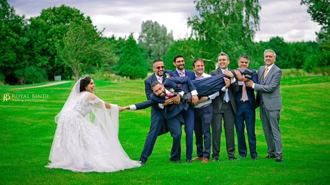 A bride in a white wedding dress is looking at a group of men in suits carrying another man. the bride is holding out her hand as if to catch the man. the men are laughing and having fun. the scene is outdoors in a green grassy area.