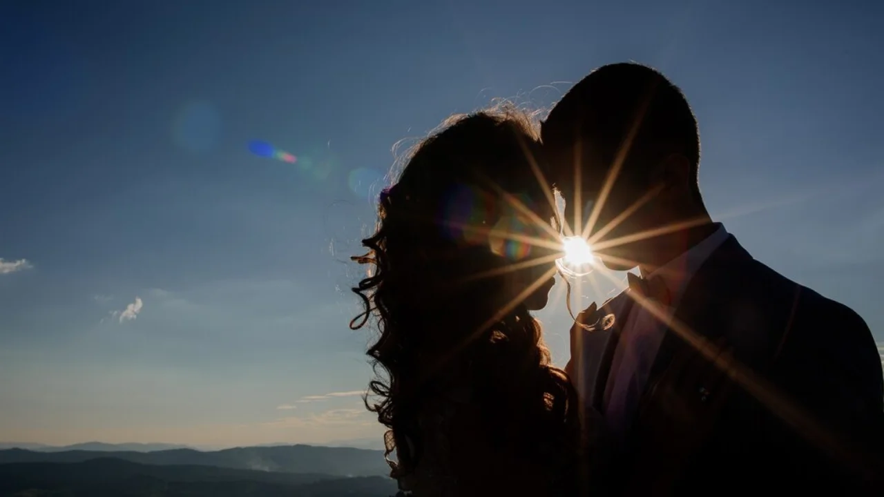 A silhouette of a couple embracing against the setting sun. The couple's heads are tilted towards each other, and the sun is shining behind them. They look very much in love.