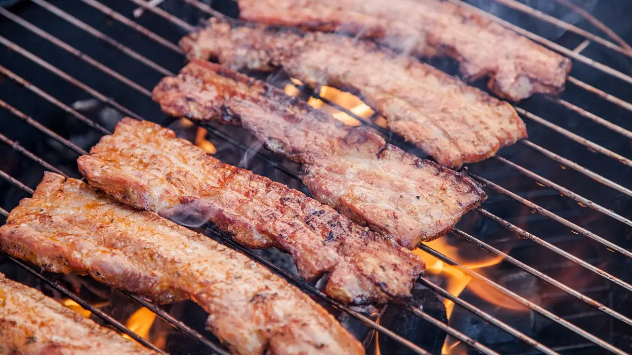 Strips of bacon sizzling on a grill over hot coals, with smoke rising from the meat.