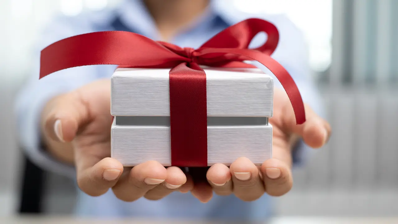 A pair of hands holding a white gift box tied with a red ribbon.