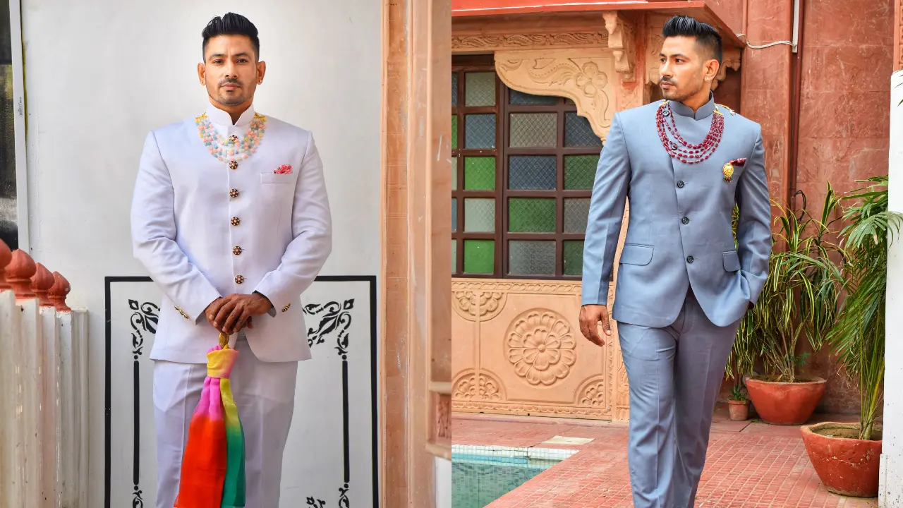 Two men in suits Alt: Two men are wearing suits, one in white and one in blue. The man in white is holding a colorful scarf. The man in blue is looking to the left. They are both standing in front of a wall with an ornate door.