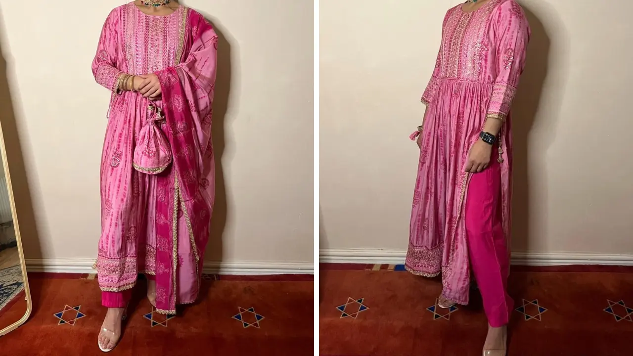 A pink salwar kameez set with a long, embroidered kameez, matching trousers, and a dupatta. The kameez has a round neckline and long sleeves. The set is paired with silver jewelry and heels.