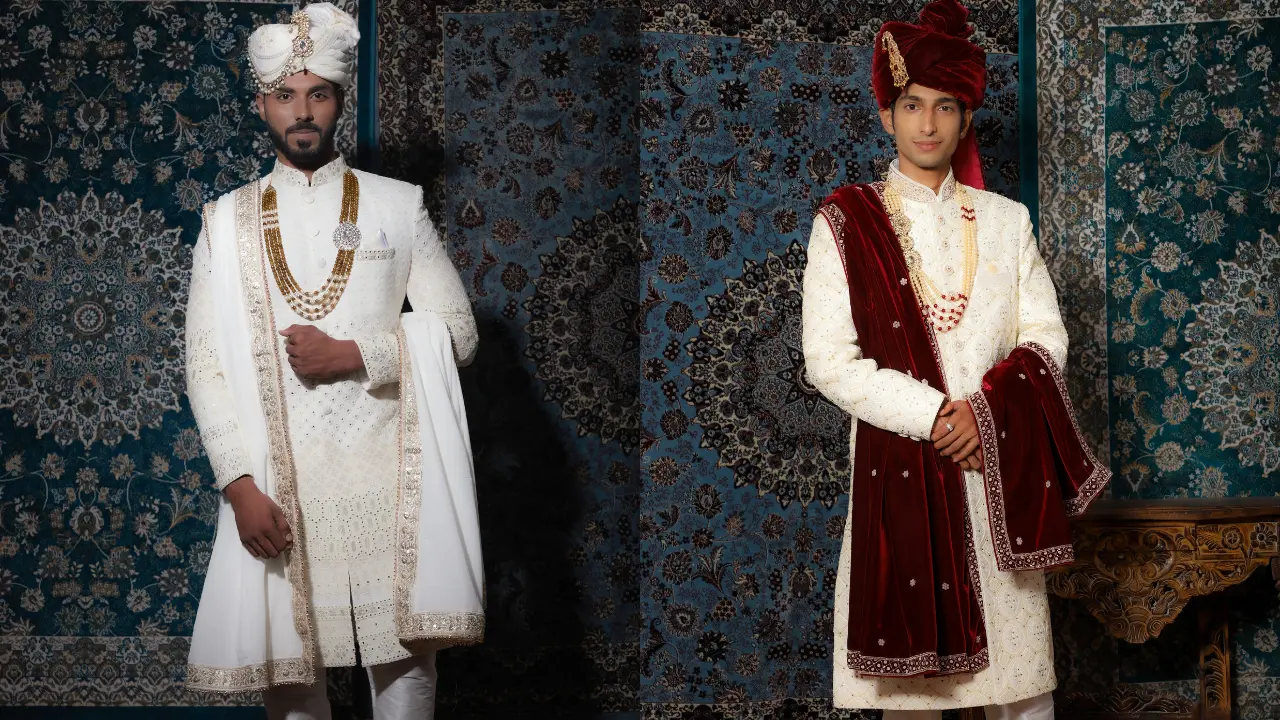 Two men wearing traditional Indian wedding attire stand in front of a colorful patterned background. The man on the left is wearing a white sherwani and a white turban. The man on the right is wearing a white sherwani and a maroon turban with a maroon shawl. Both men are smiling. The background is a blue and white patterned carpet.