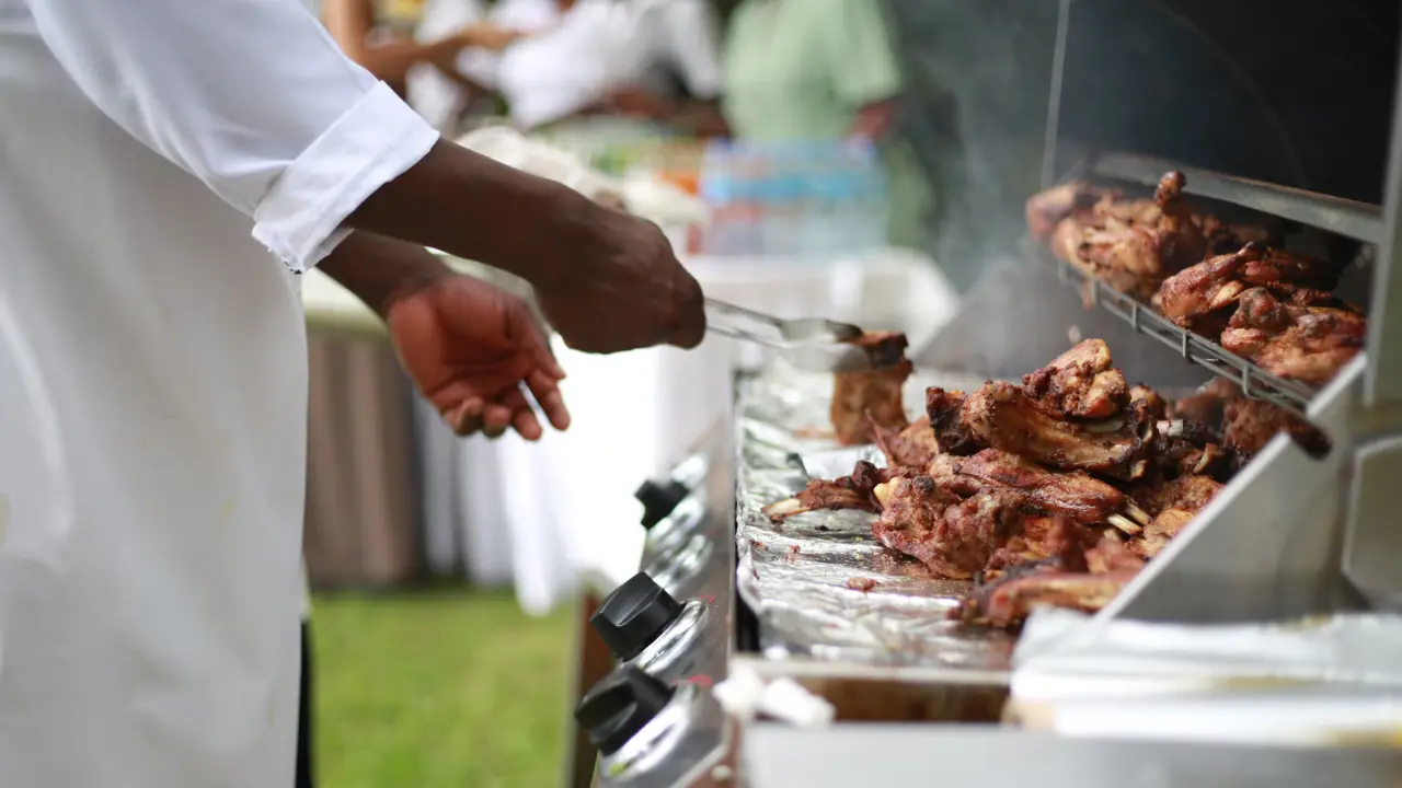 Close-up of a chef grilling meat on an outdoor barbecue grill at a live cooking station, with smoke rising from the grill.