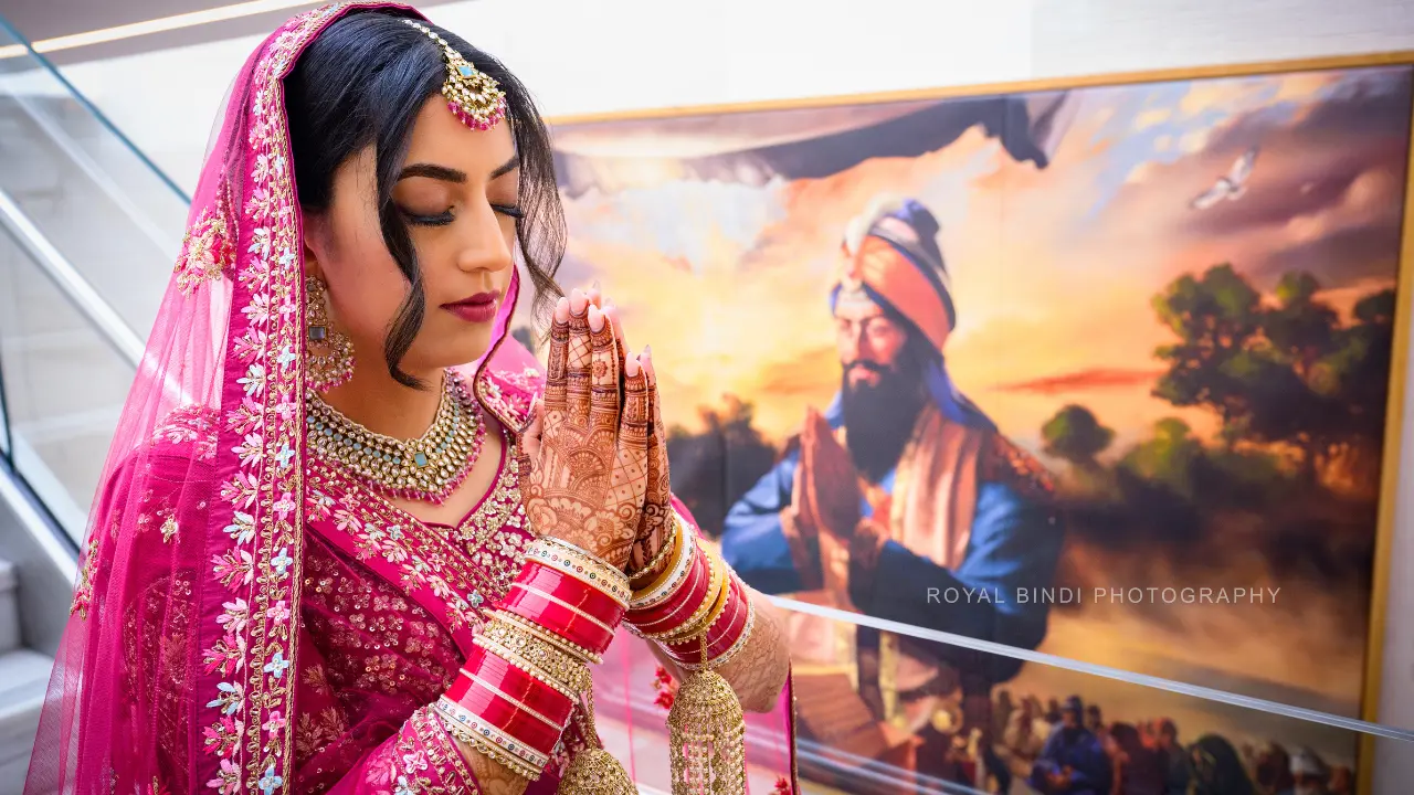 A bride in a pink and gold lengha and dupatta stands in front of a painting of a Sikh man in prayer. She has her hands clasped together in a prayerful gesture, and a large bindi on her forehead. The bride is wearing a lot of jewelry, including bangles, earrings, and a necklace.