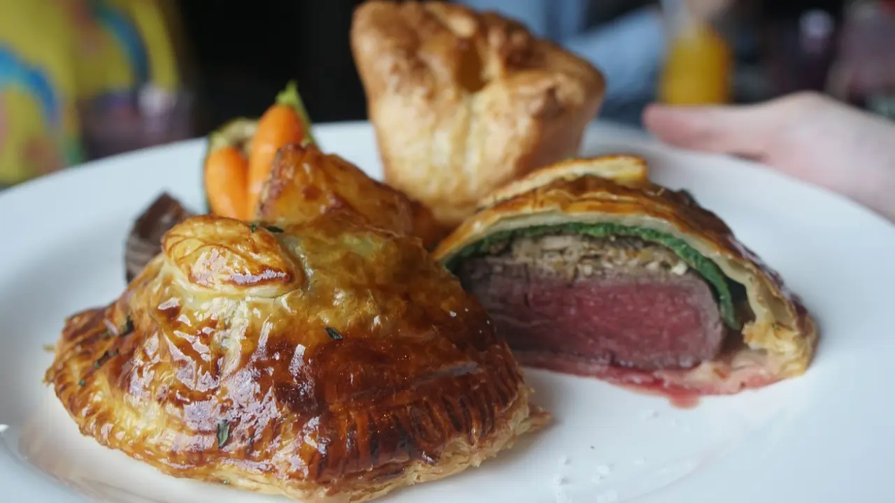 A plate with a beautifully presented Beef Wellington, sliced open to reveal the tender meat and mushroom filling. The dish is accompanied by roasted vegetables and a Yorkshire pudding.