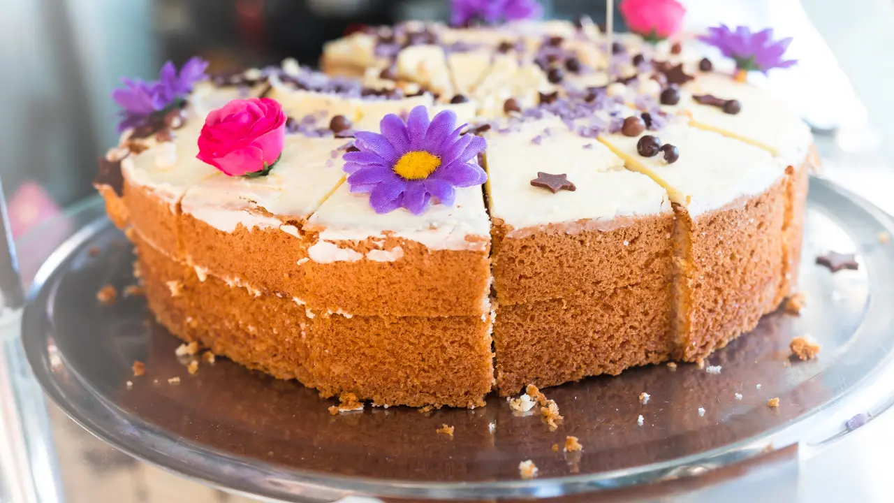 A round cake with white frosting and colorful decorations, including flowers and sprinkles. The cake is sliced into wedges and displayed on a silver tray.