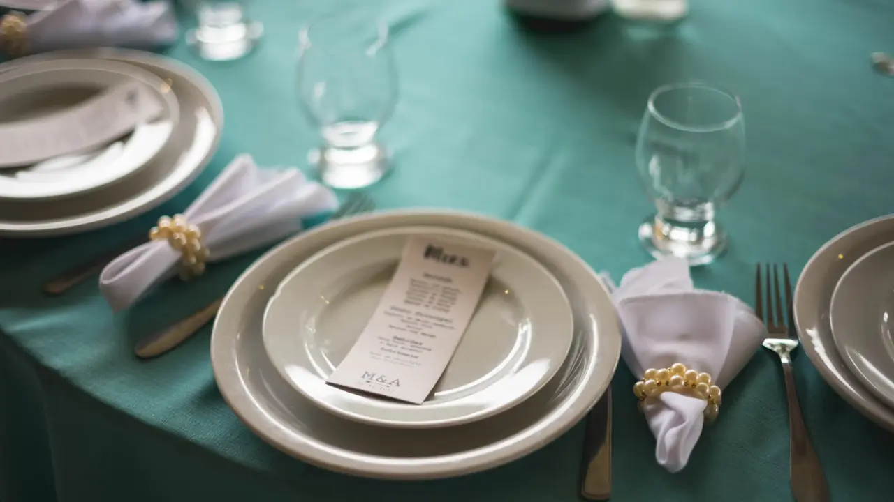 A close-up of a beautifully set table with white plates, glasses, silverware, and napkins adorned with pearl napkin rings. A menu card rests on one of the plates. The table is covered with a vibrant turquoise tablecloth.