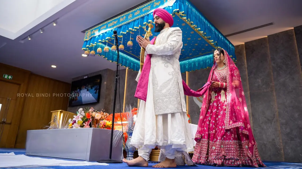 A Sikh bride and groom are standing under a canopy during a wedding ceremony. The groom is wearing a white sherwani and the bride is wearing a pink lengha.