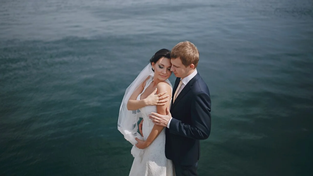 A newly married couple standing by a lake, smiling and holding each other. The bride is wearing a white wedding dress, and the groom is wearing a black suit. They are both looking at each other with love and joy.