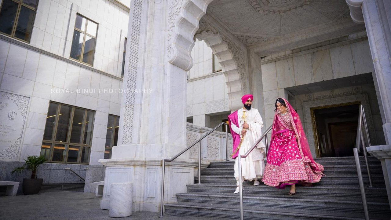 A Sikh couple in traditional wedding attire, the man wearing a white turban and the woman wearing a pink dress and veil, walk up the steps of a gurdwara, a Sikh temple.