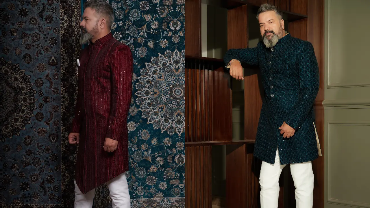 Two men in traditional Indian clothing stand in front of a wooden wall. The man on the left wears a long burgundy jacket and white pants. The man on the right wears a long teal jacket and white pants. Both men look confident and stylish. This image is about traditional clothing and style.