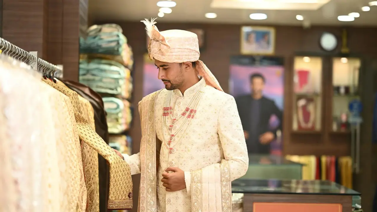 A man dressed in a white sherwani and a pink turban is browsing a display of garments at a shop. He is smiling as he looks at the clothes. The shop is decorated with brown wood paneling and shelves lined with fabric.