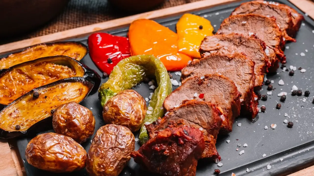 A platter of grilled meat sliced and served with roasted vegetables like potatoes, eggplant, and peppers.