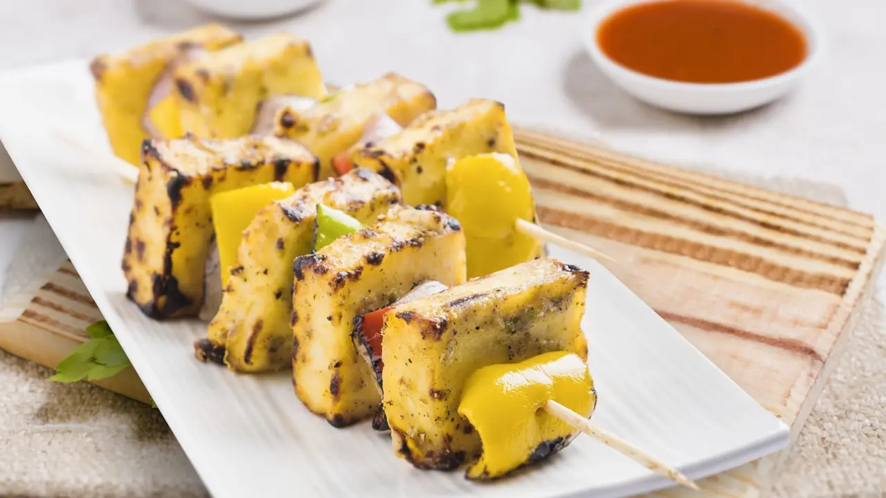 A plate of grilled paneer tikka skewers with bell peppers, served with a bowl of red dipping sauce.