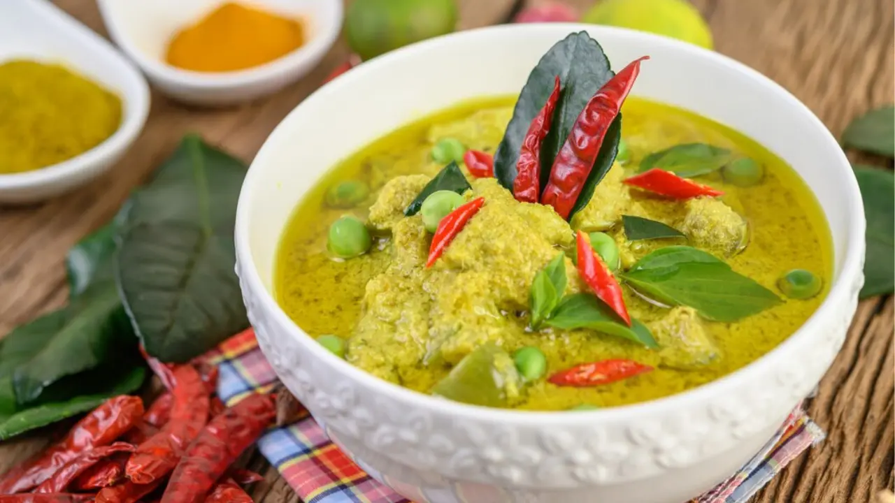 A bowl of green curry with chunks of meat, peas, and red chili peppers, garnished with fresh herbs.