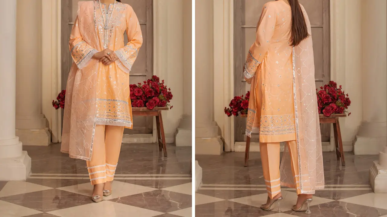 A woman wearing a peach and silver embroidered suit. She is standing in a room with white walls and red flowers. She is looking at the camera.