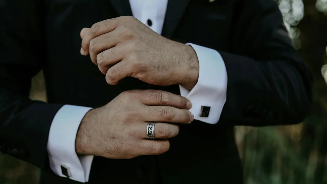 A close-up photo of a groom's hand holding a gold wedding ring.