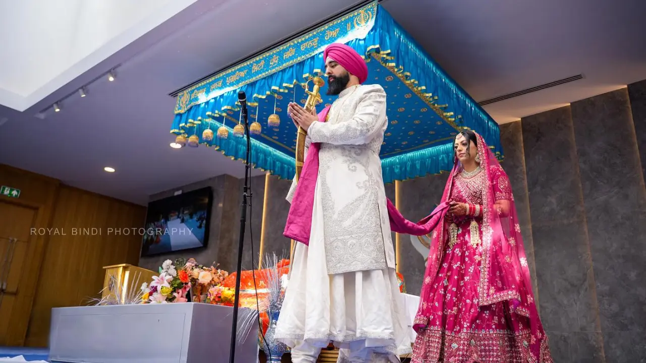 A Sikh couple are getting married in a beautiful ceremony. The bride is wearing a pink and gold dress with a red veil, and the groom is wearing a white suit and turban. The ceremony is taking place in a temple, and there are floral decorations and lights.