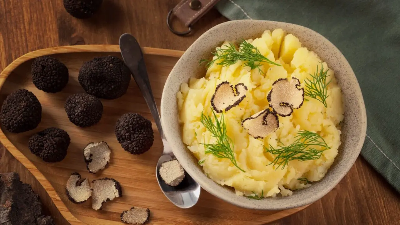 A bowl of creamy mashed potatoes topped with fresh dill and black truffle shavings. A wooden tray with whole black truffles is in the background.