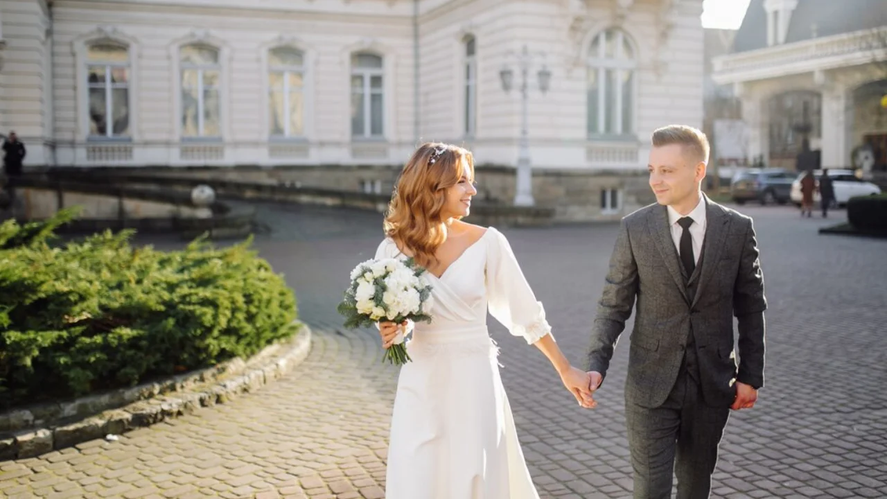 A bride and groom holding hands and walking down a cobblestone street. The bride is wearing a white dress and the groom is wearing a grey suit. They are smiling and looking at each other.