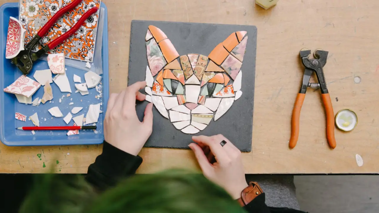 A person's hands carefully placing small pieces of ceramic tile to form a mosaic of a cat's face.