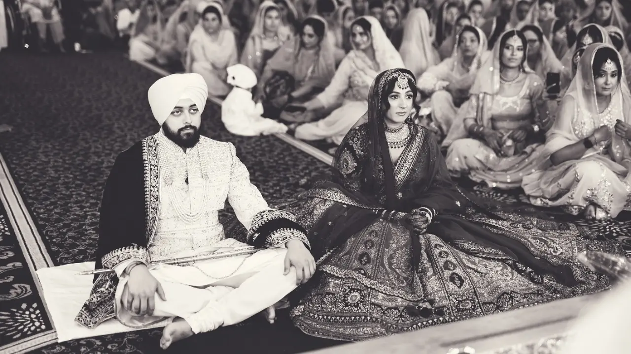 A black and white photo of a newly married couple sitting on a carpet in a traditional setting. The groom is wearing a turban and a long, white robe, while the bride is wearing a beautiful, ornate gown and a veil. They are surrounded by other people seated in the background.