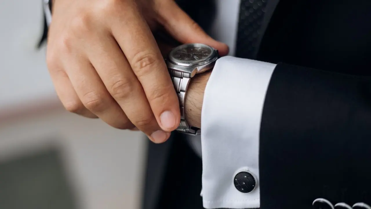 A man's hand adjusting a silver wristwatch, with cufflinks visible on his shirt cuff.