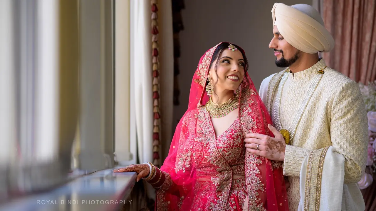 A Sikh bride in a vibrant red lehenga and jewellery smiling warmly at her groom in ivory sherwani and turban, captured beautifully by Royal Bindi Photography near a window.