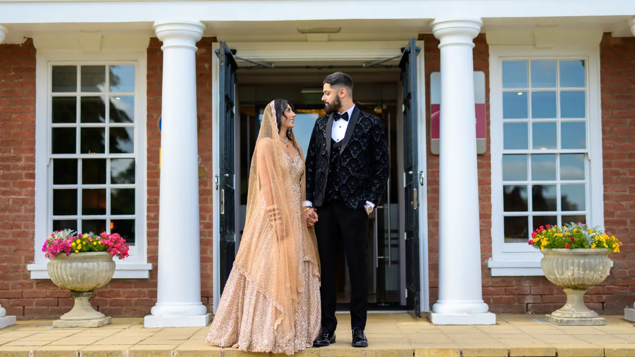 A bride in a golden traditional outfit and a groom in a black tuxedo standing hand in hand at the elegant entrance of the National Motorcycle Museum Birmingham.