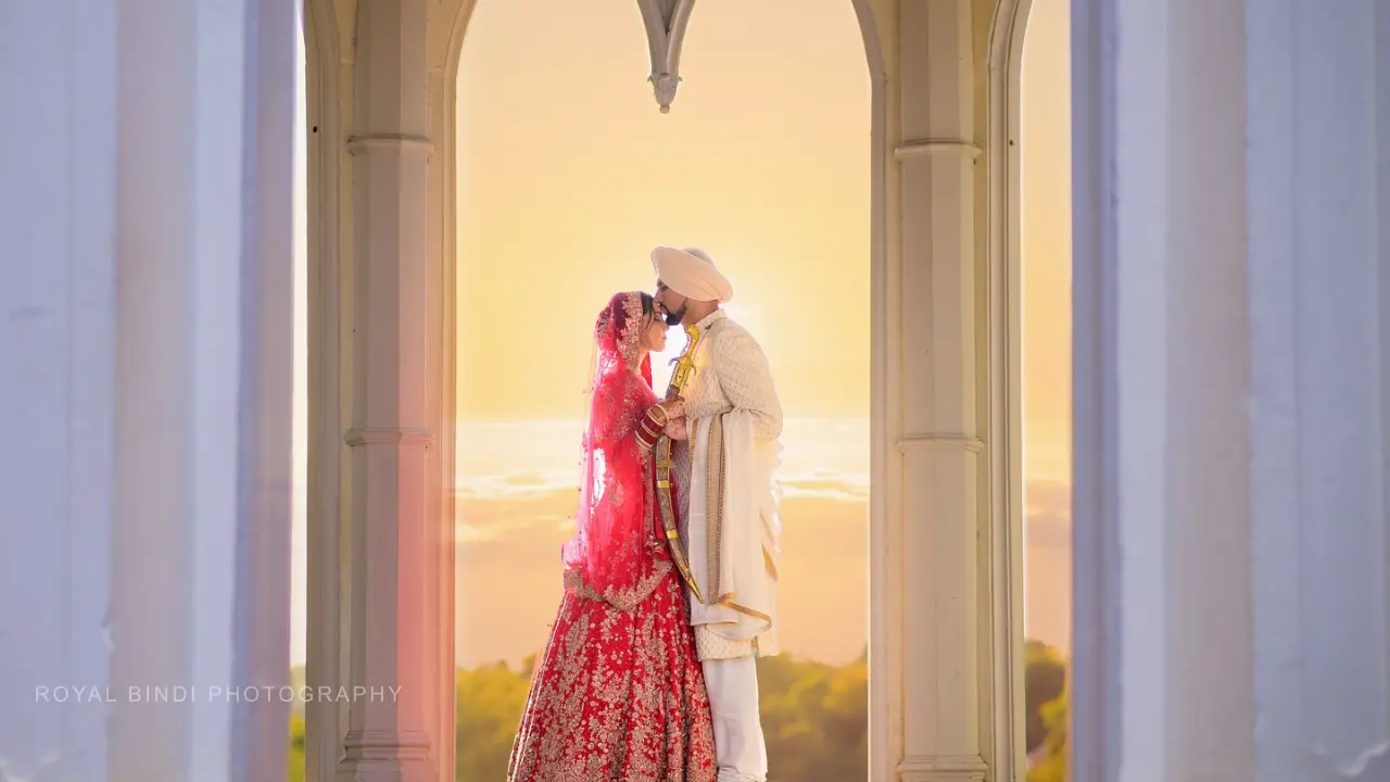 A Sikh bride and groom share a romantic moment under an archway at sunset, wearing traditional wedding attire, captured by Royal Bindi Photography.