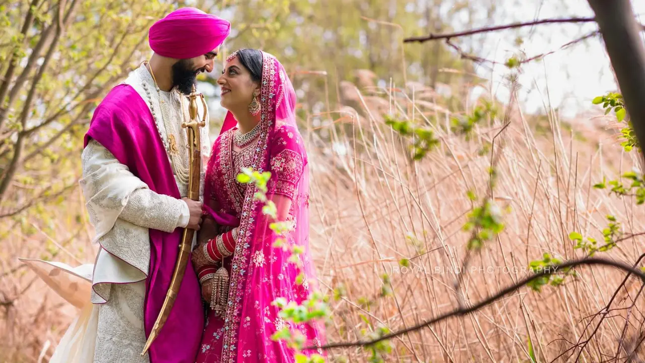 A Sikh groom and bride in traditional pink and ivory wedding attire, sharing a loving moment in a natural outdoor setting with tall grasses and greenery.